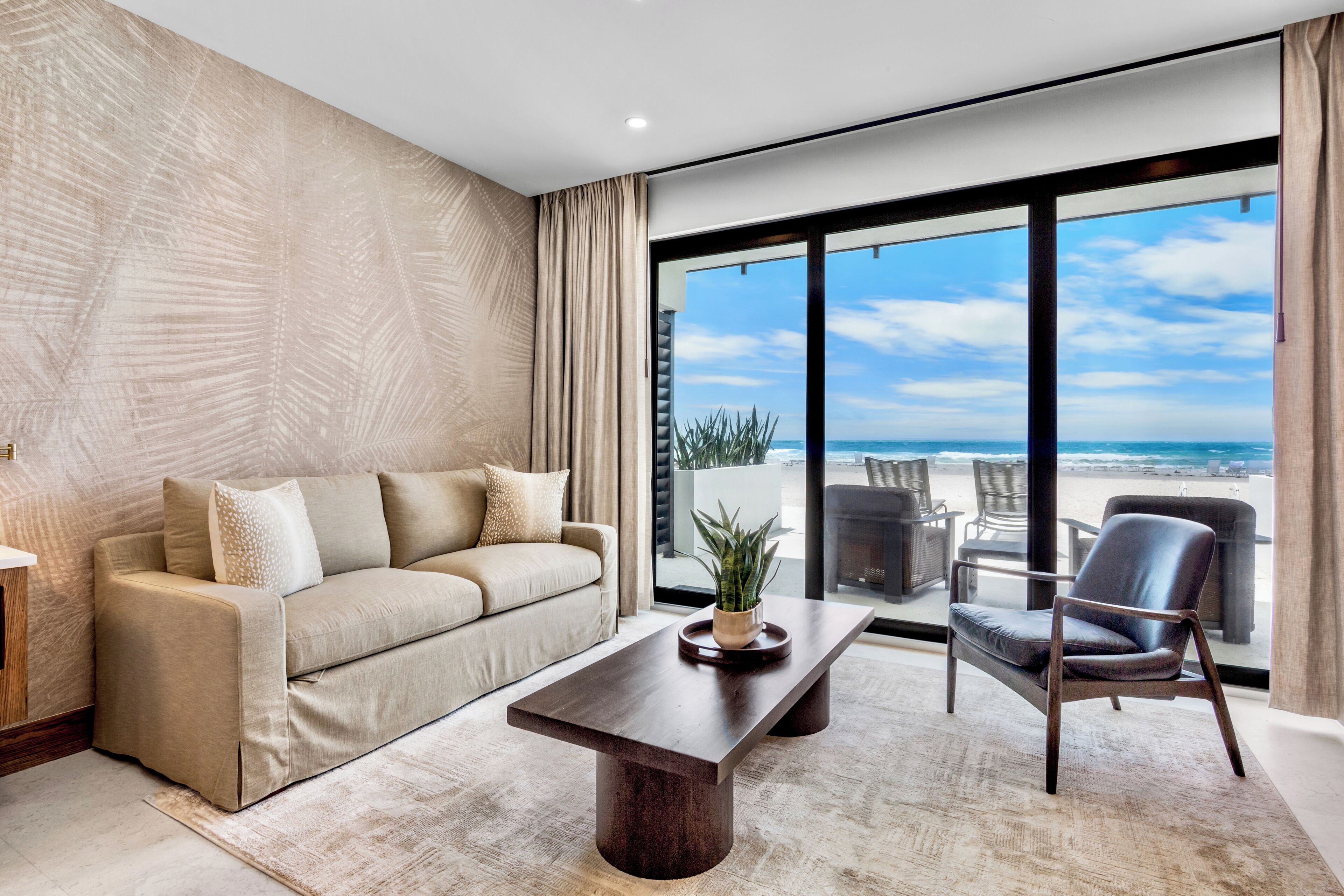 A modern living room with a sofa, chairs, a coffee table, and a view of the beach.