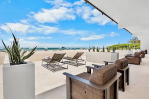 Eine Terrasse am Strand mit Korbmöbeln, einem weißen Pflanzgefäß mit Pflanze und Blick auf den Ozean.