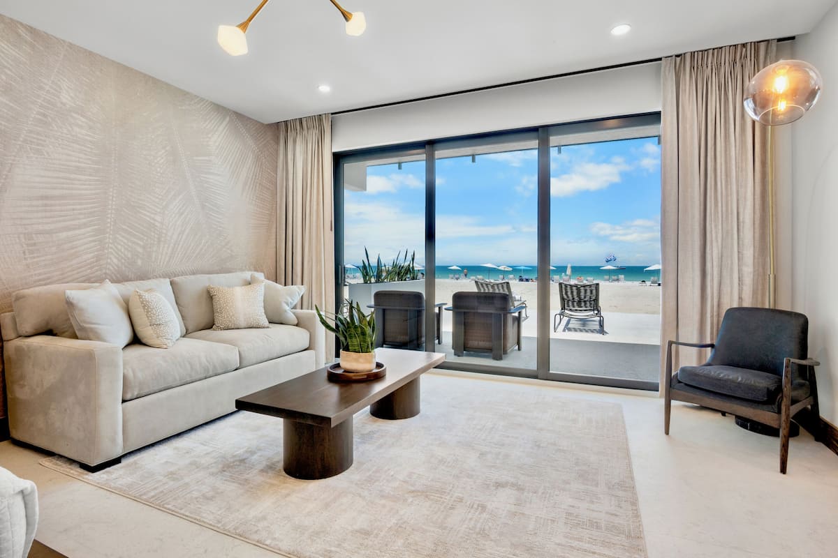 A modern living room with a sofa, a coffee table, and a chair, a large window with a view of the beach, and a balcony with outdoor furniture.