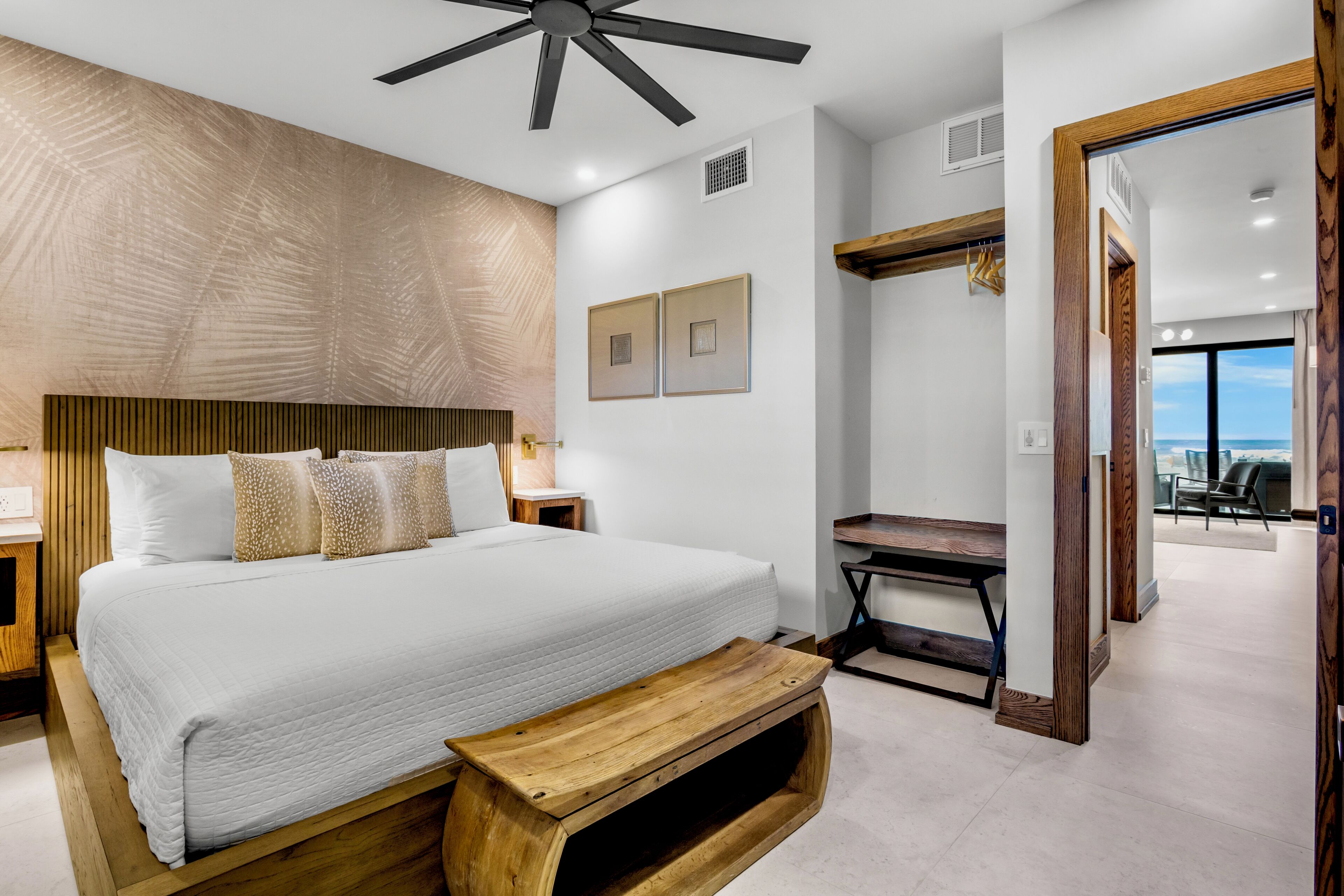 A modern bedroom with a large bed, a wooden bench, a ceiling fan, and a view of the ocean through a large window.