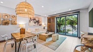 A modern living room with a wooden dining table, white chairs, a sofa, a coffee table, and a large woven pendant light. There are wall art pieces and a large window with a view of palm trees. - Bali Hai Beachfront Resort and Spa (Holmes Beach)