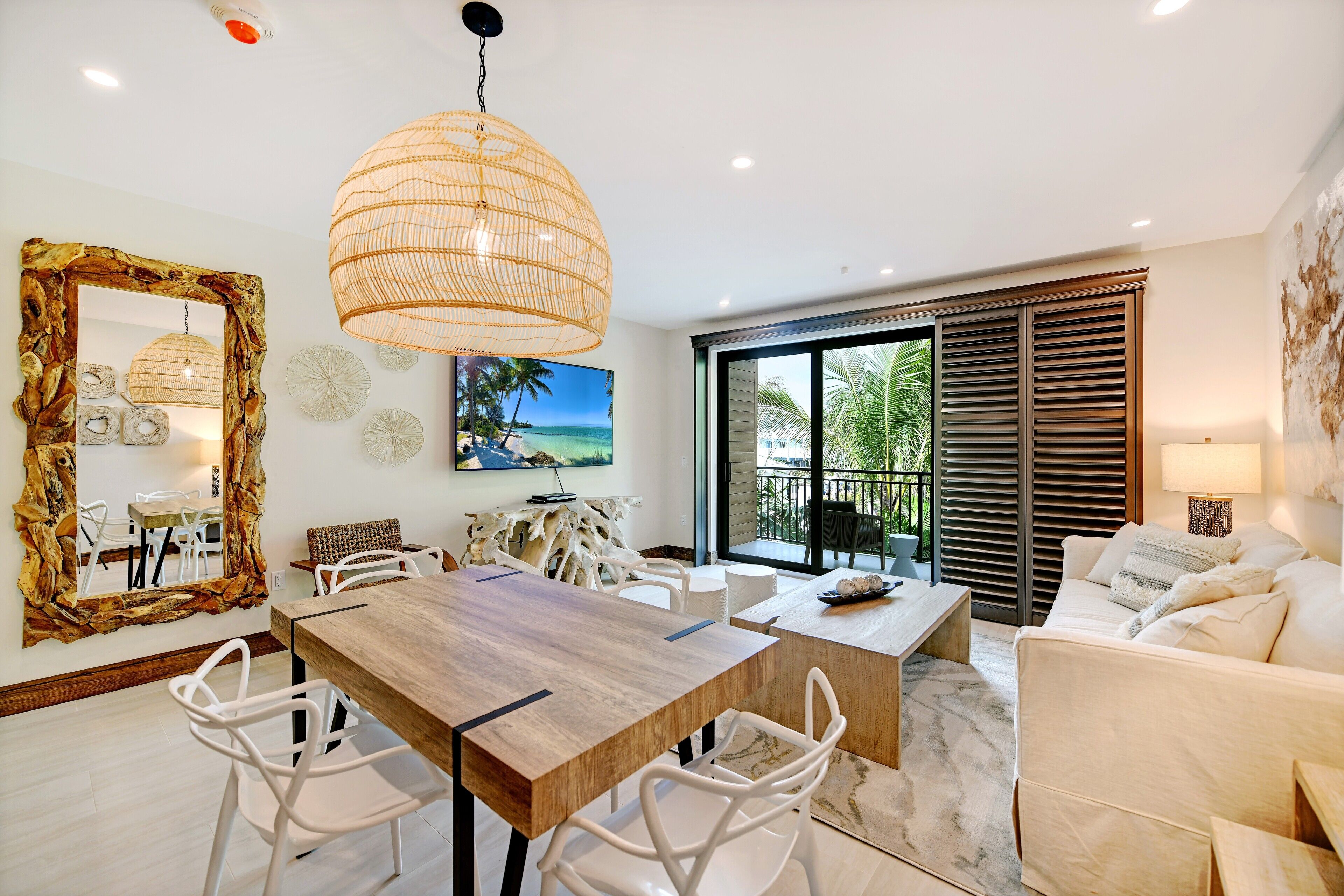 A modern living room with a wooden dining table, white chairs, a large mirror, and a view of the ocean through large windows.