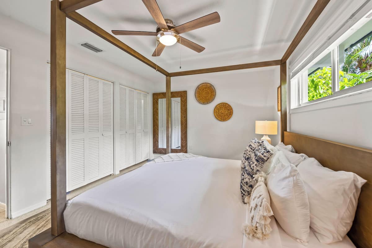 A bedroom with a large bed, a ceiling fan, a window with a view of greenery, and a door with white shutters.
