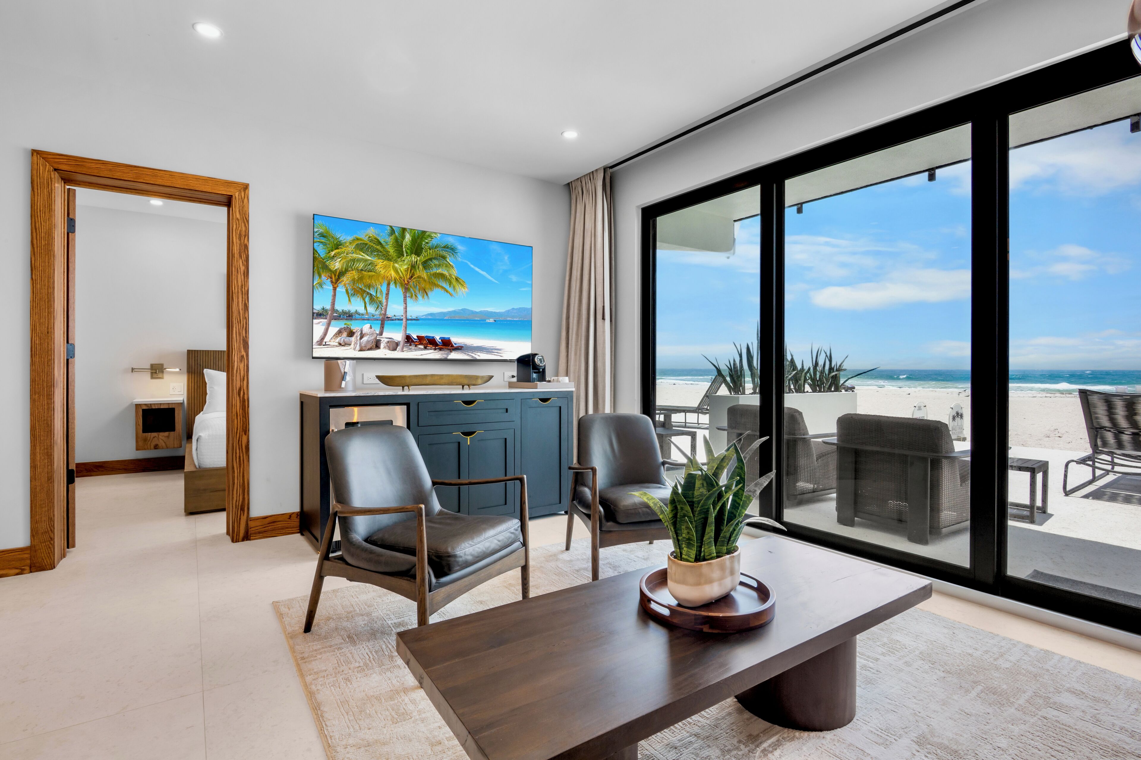 A modern hotel room with a large window offering a beach view, a dining area with a table and chairs, and a seating area with a view of the ocean.