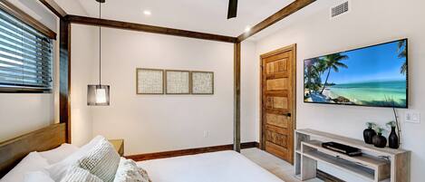 Un dormitorio con una cama, un ventilador de techo, un televisor sobre un mueble de madera y una puerta con panel de madera.