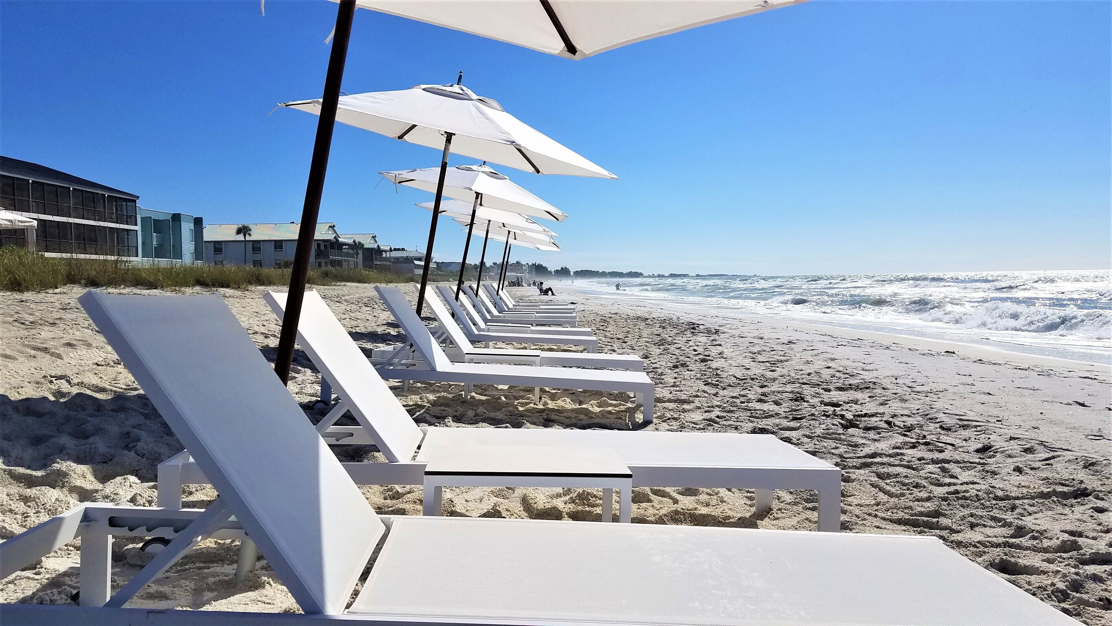 A beach with white lounge chairs and umbrellas, a sandy shore, and a building in the background.