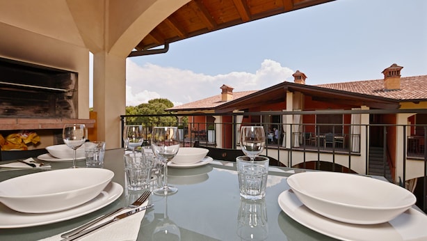 A dining table set with white plates, bowls, cutlery, and wine glasses, with a view of a building and trees in the background. - Appartamento Romantica (Manerba del Garda)