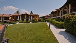 A courtyard with a well-maintained lawn, a paved walkway, and a row of buildings with arched windows and balconies. - Appartamento Romantica (Manerba del Garda)