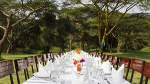 Una mesa dispuesta al aire libre con manteles blancos, sillas de madera y un centro de mesa floral.