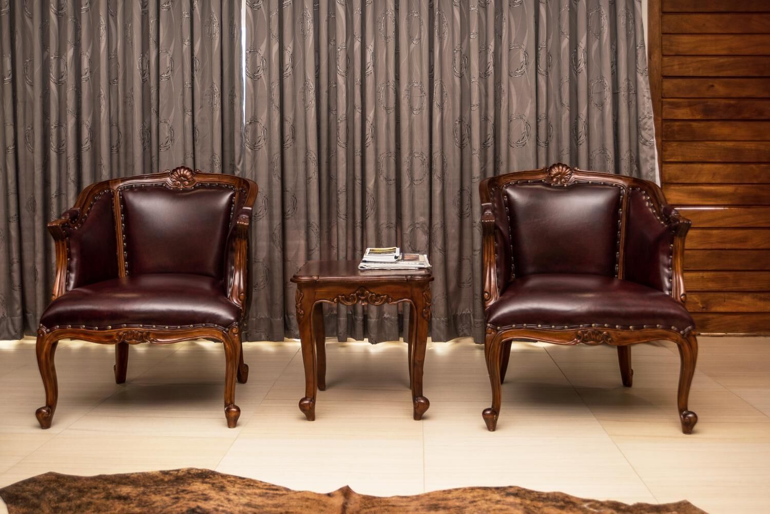 Dos sillones de cuero con patas de madera y una pequeña mesa de madera con un libro en el centro, frente a cortinas estampadas y una pared de madera.