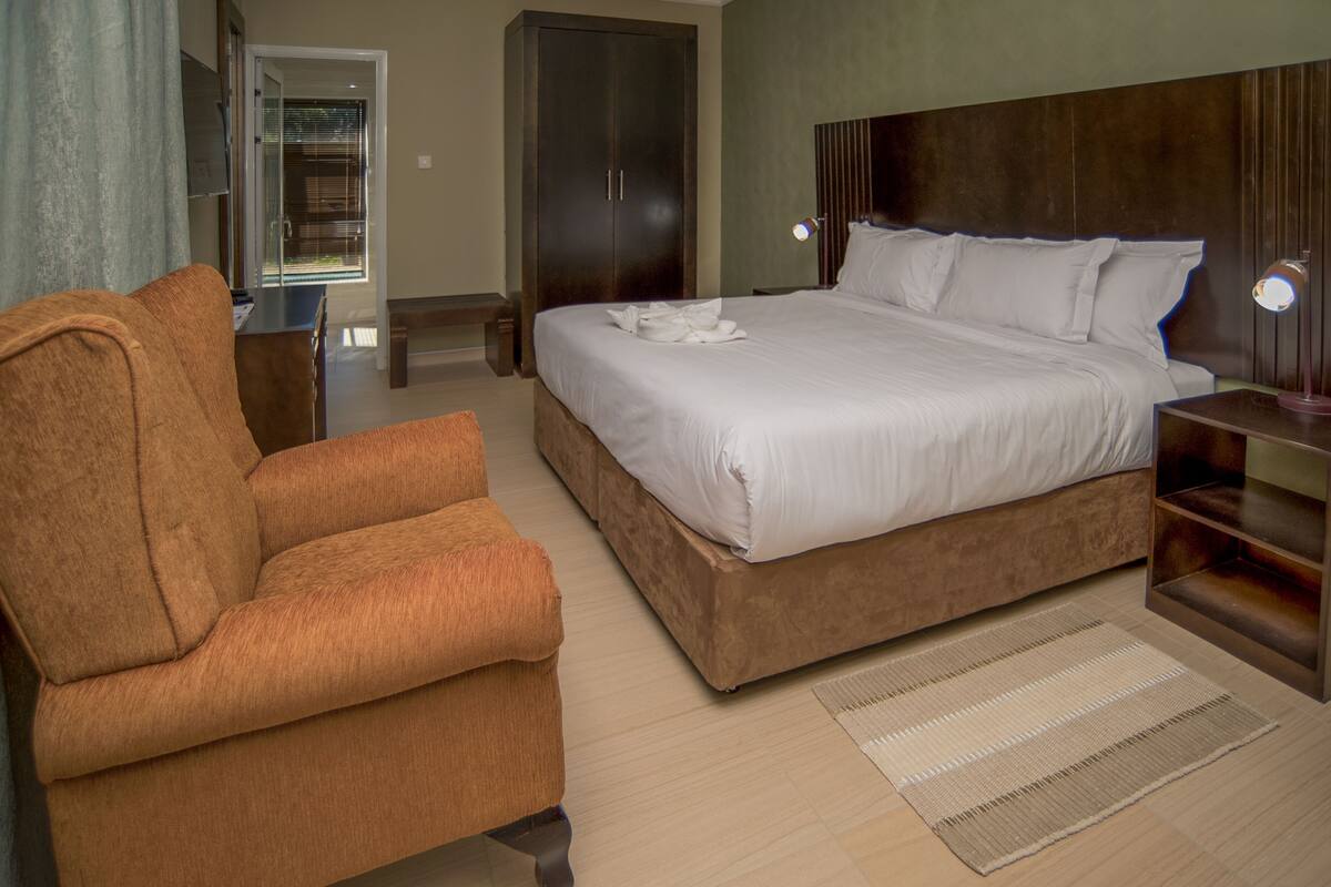 A bedroom with a large bed, a brown armchair, a wooden wardrobe, and a bedside table with a lamp.