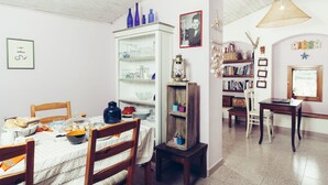 A dining area with a wooden table set for four, surrounded by chairs. A white cabinet with glassware is to the left, and a bookshelf with books and decorative items is to the right. - B&B Blu Bosa (Bosa)