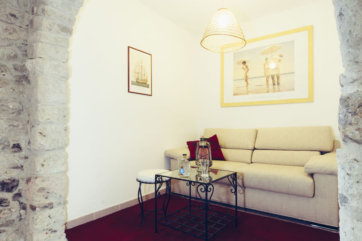 Una habitación con pared de piedra, un sofá color beige, una mesa de centro de vidrio con una botella y un farol, y cuadros en la pared.