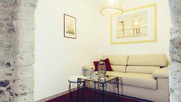 Une pièce avec un mur en pierre, un canapé beige, une table basse en verre sur laquelle se trouvent une bouteille et une lanterne, et des tableaux encadrés accrochés au mur.