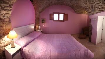 A bedroom with a stone wall, a bed with a pink bedspread, a nightstand with a lamp, and a small window.