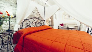 A room with a bed under a canopy, a stone wall, a small table with a lamp, and a sofa in the background. - B&B Blu Bosa (Bosa)