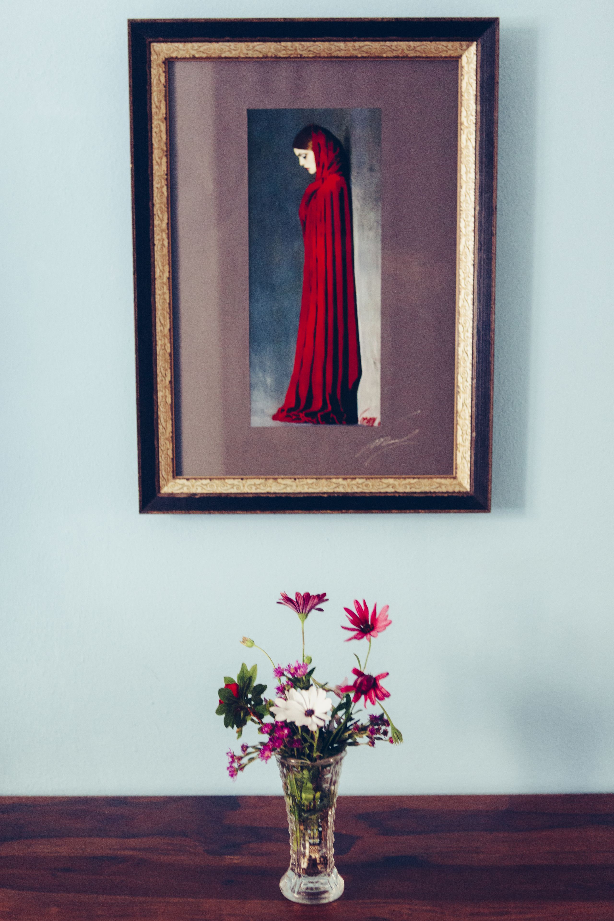 A framed artwork of a figure in a red cloak hanging on a wall, with a vase of flowers below it.