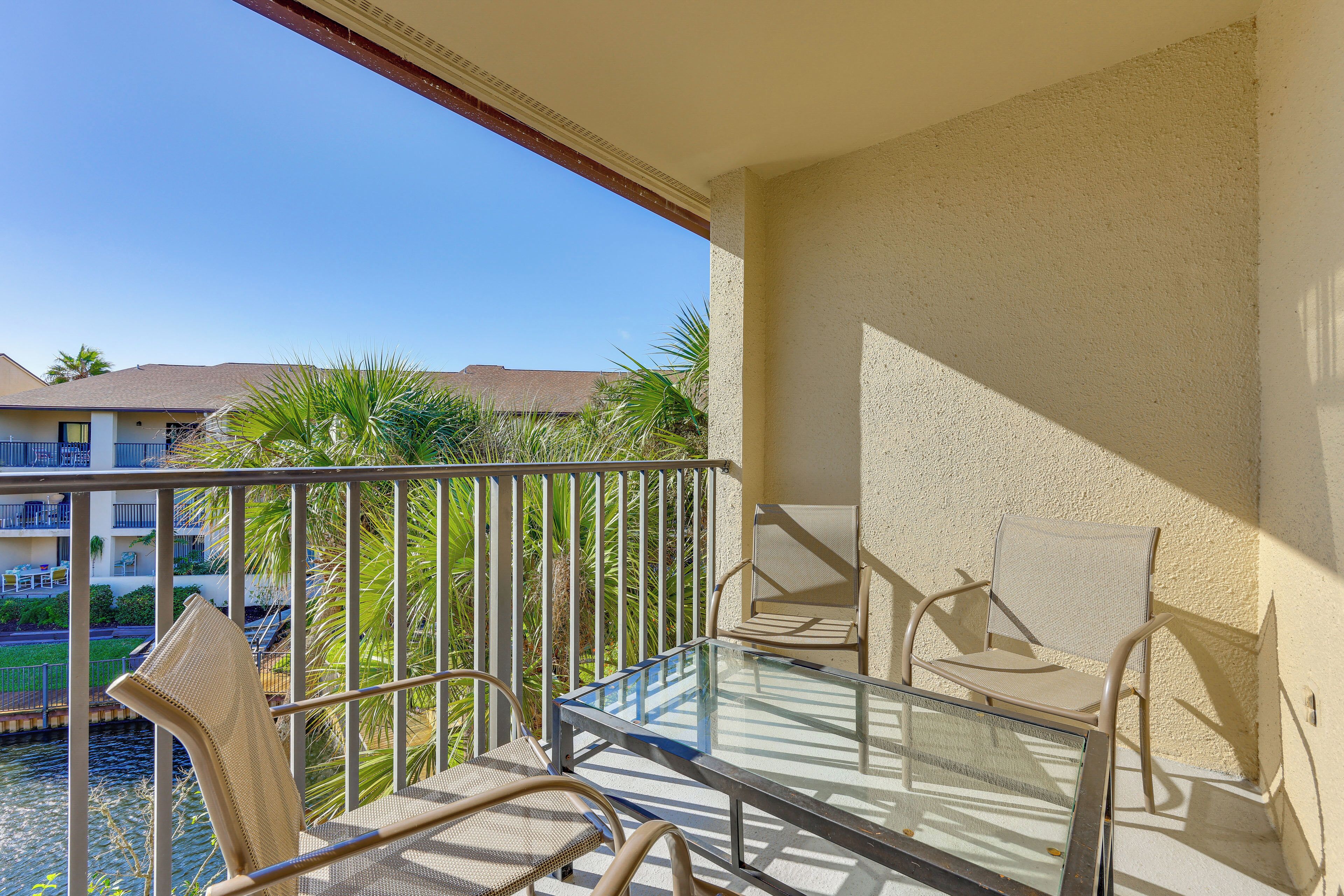 Un balcon avec une table en verre et deux chaises, donnant sur une piscine et des immeubles.