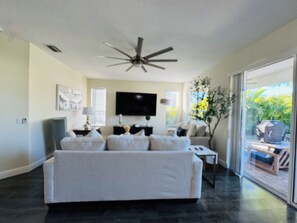 A modern living room with a large sofa, a flat-screen TV, a ceiling fan, and a view of the outdoors through a sliding glass door.