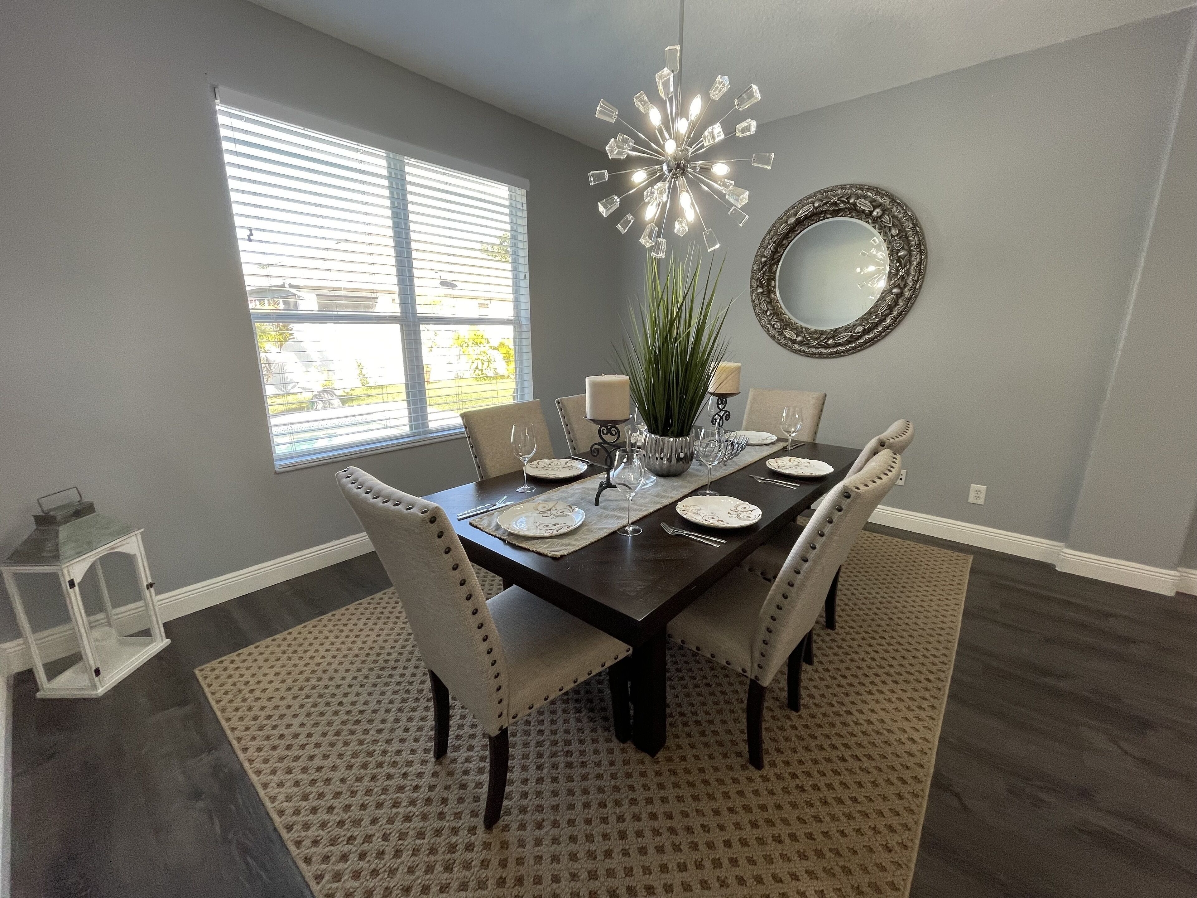 A dining room with a dark wooden table set for six, surrounded by six chairs. There is a large mirror on the wall, a chandelier, and a window with blinds.