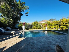 A swimming pool with lounge chairs, surrounded by lush greenery and a clear blue sky.