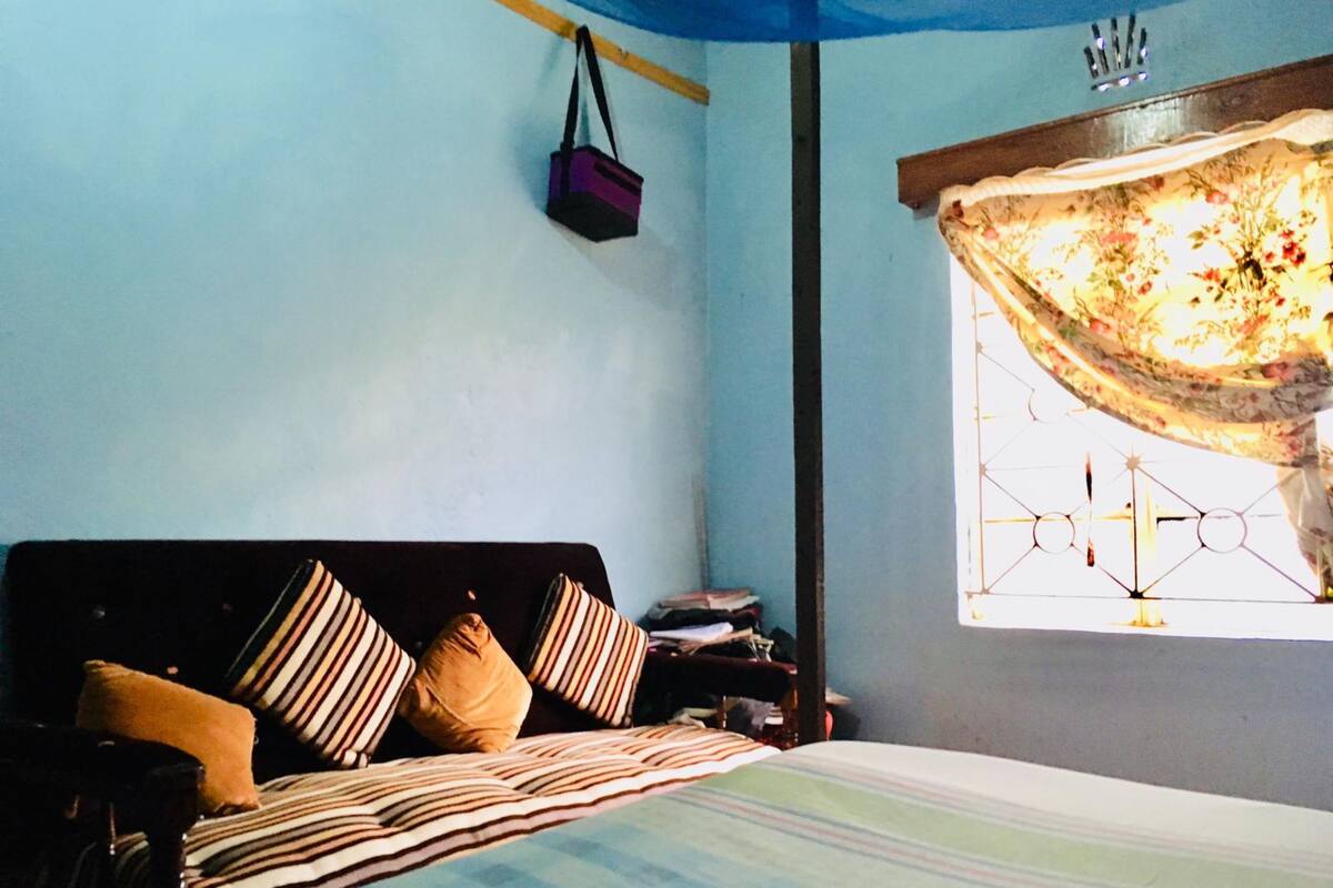 A bedroom with a bed, pillows, a nightstand, a window with a curtain, and a hanging bag.