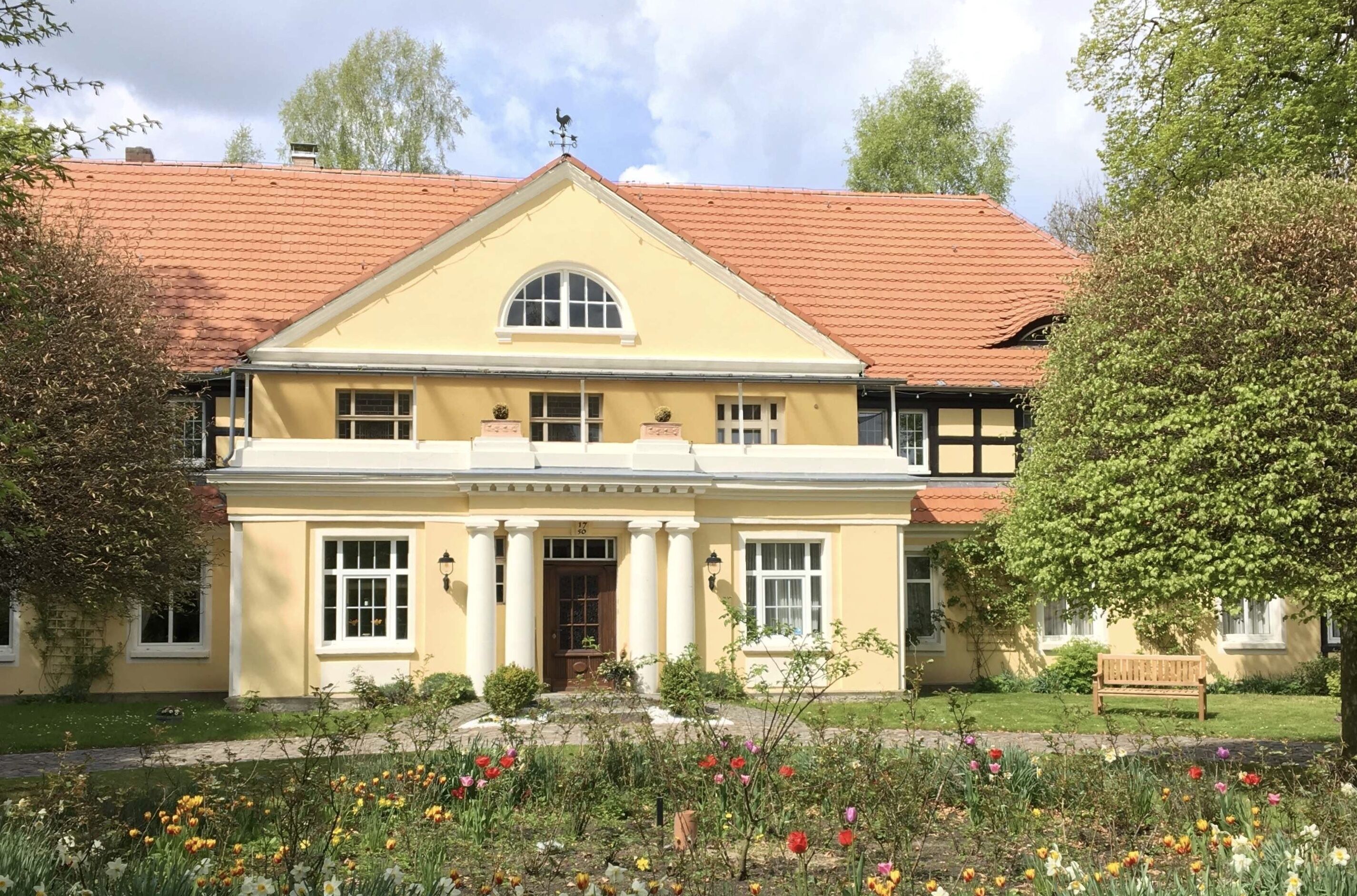 A two-story building with a yellow facade, red-tiled roof, and a symmetrical front entrance with columns.