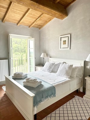 A bedroom with a bed, a nightstand, a lamp, a framed picture, and a window with a view of greenery. - Luxury 6-bed Tuscan Villa Near Lucca and Florence (Capannori)