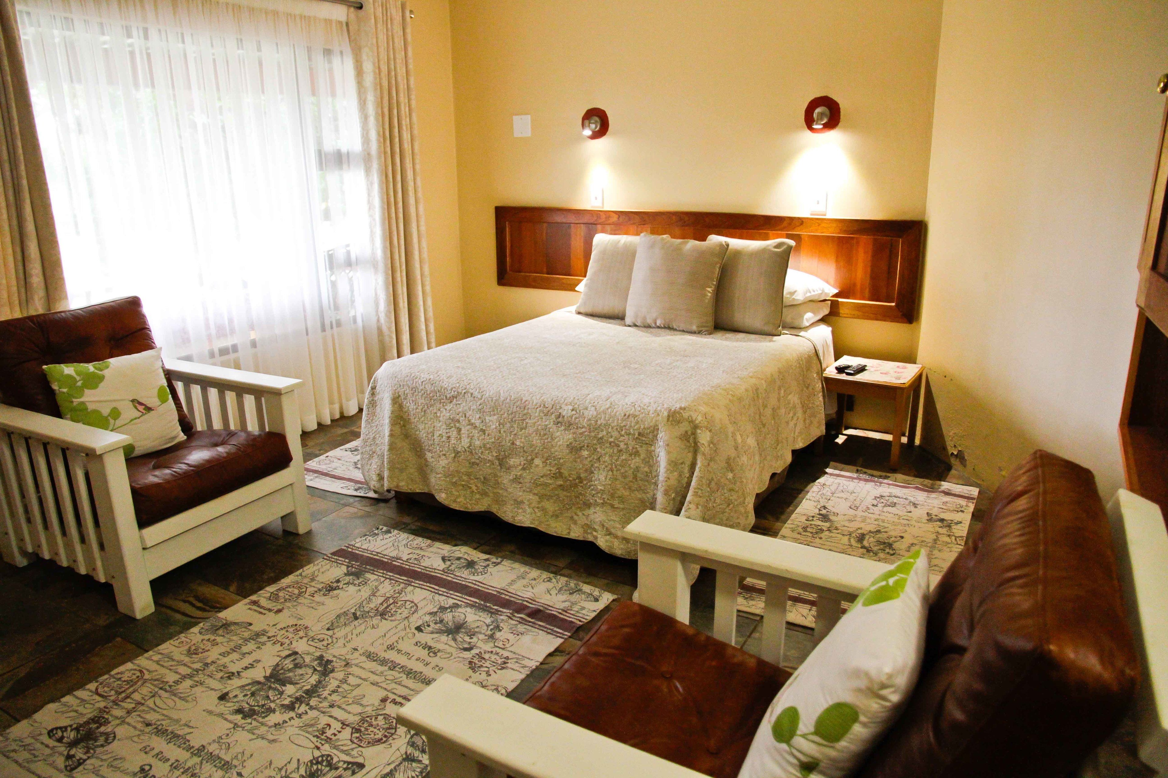 A hotel room with a large bed, two bedside tables, a nightstand, a brown leather armchair, and a white armchair with green pillows.