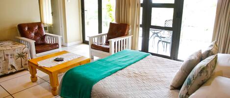 A hotel room with a bed, two armchairs, a small table, and a view through a glass door to an outdoor area.