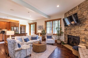 A living room with a stone fireplace, a flat-screen TV mounted on the wall, and a kitchen in the background.
