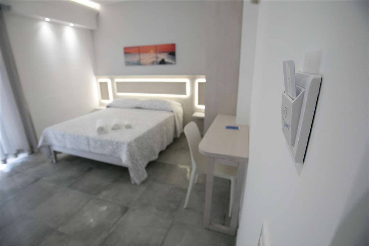 A bedroom with a bed, a desk, and a chair. There is a painting on the wall and a light fixture above the bed.