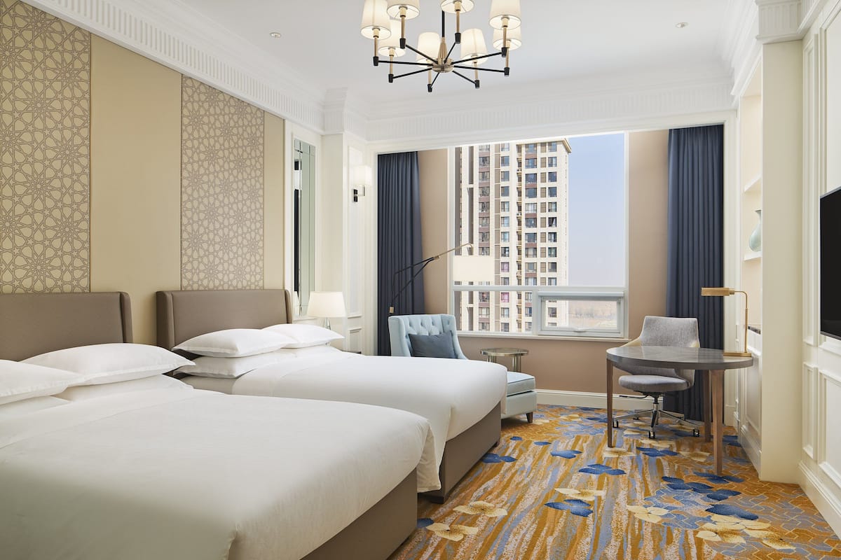 A hotel room with two beds, a small table, a chair, a TV, and a window with a city view.