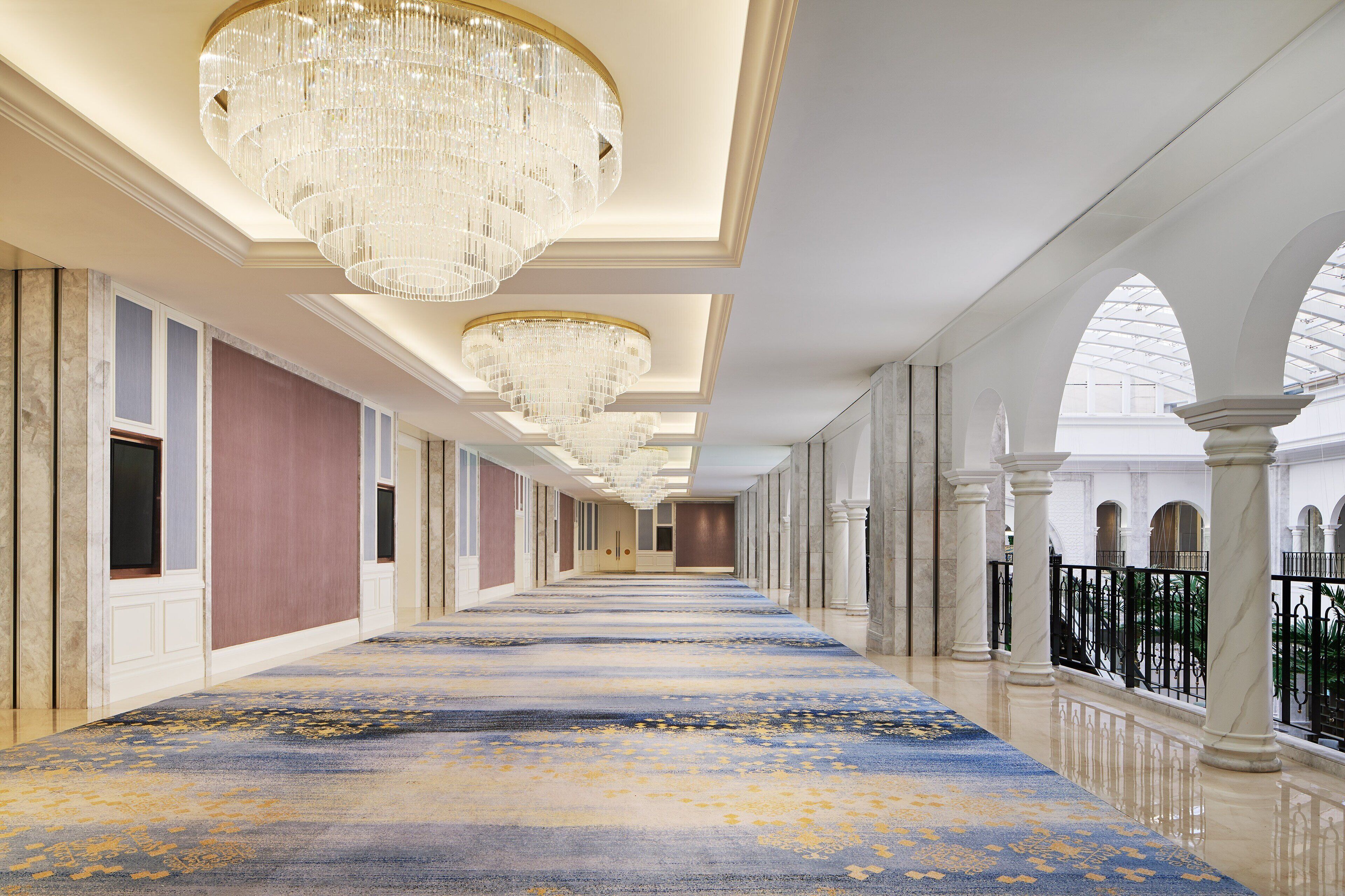 A long corridor with a blue carpet, chandeliers, and columns.