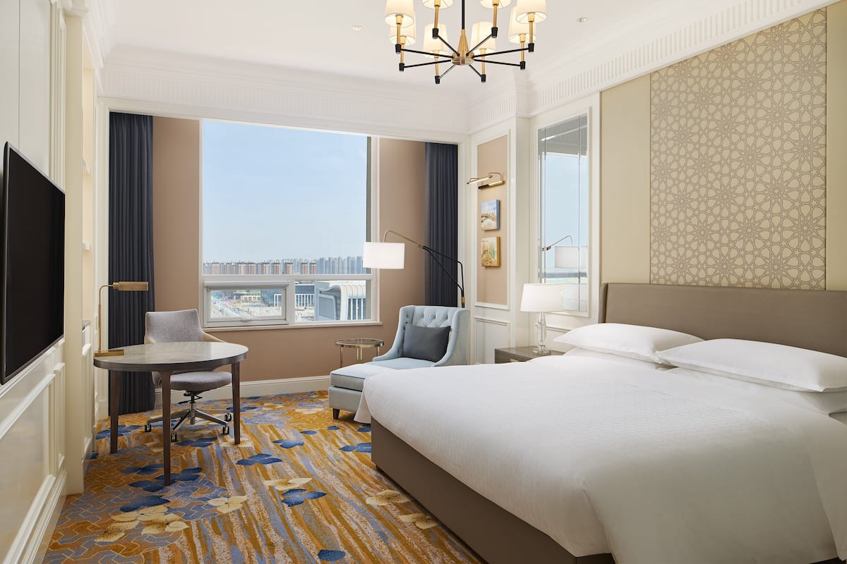 A hotel room with a large bed, a desk, a chair, a TV, and a window with a city view.