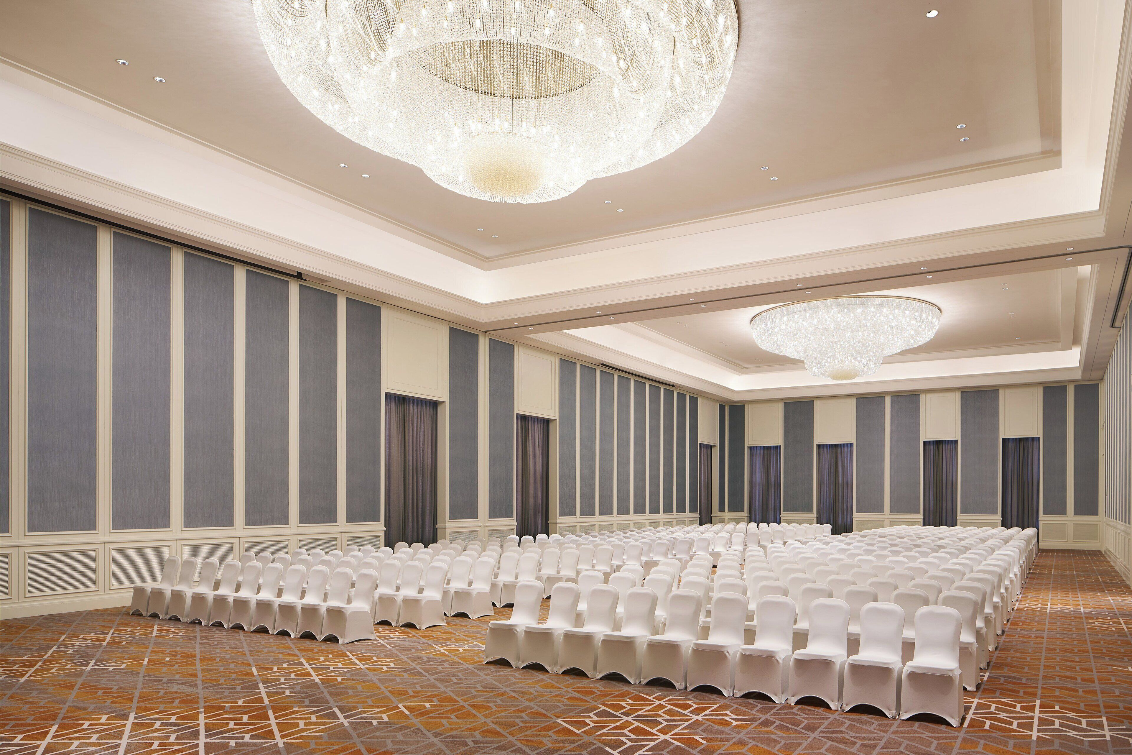 Un salón de conferencias con filas de sillas blancas, un suelo estampado y una gran lámpara de candelabro.