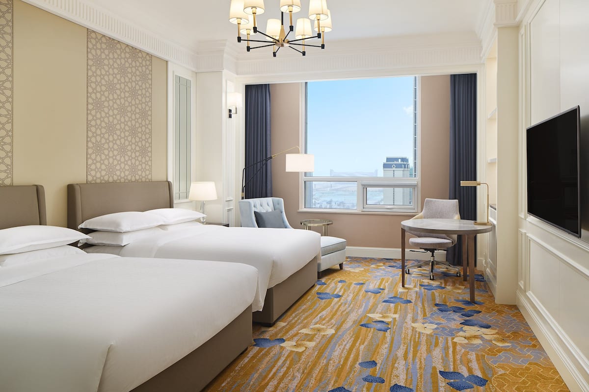 A hotel room with two beds, a small table, a chair, a TV, and a view of the city.