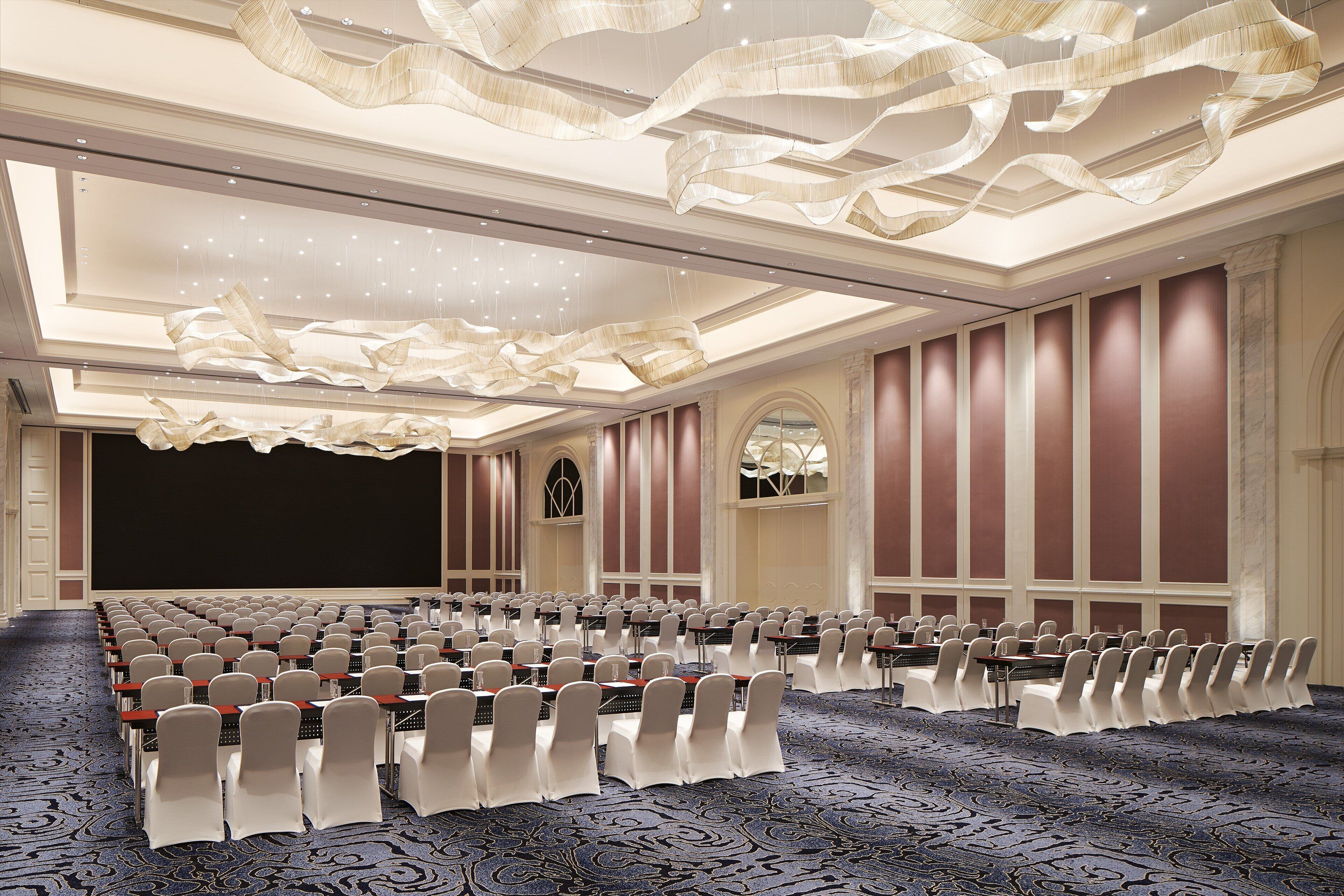 Une salle de conférence avec des rangées de tables et de chaises, un grand écran et une moquette à motifs.