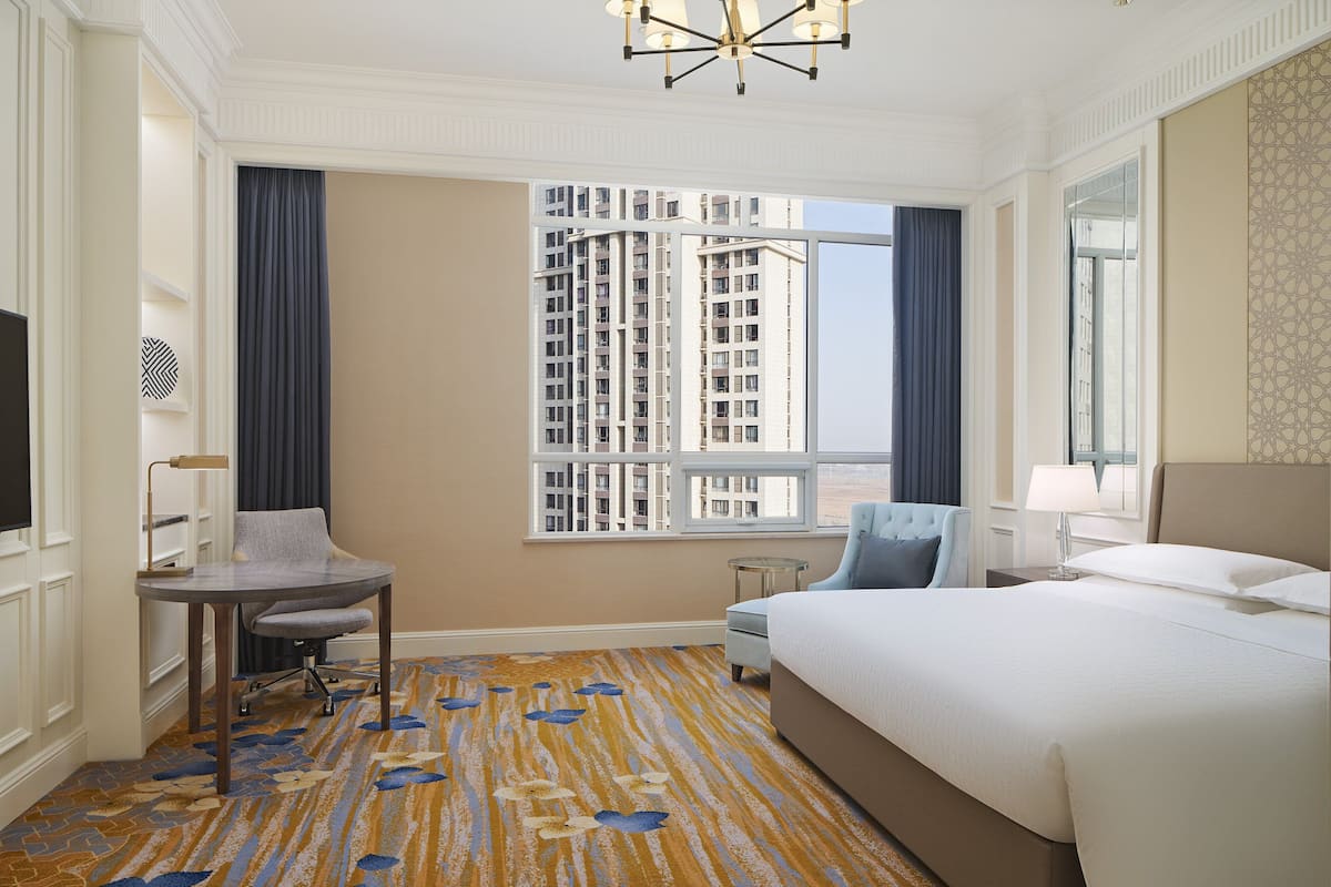 A hotel room with a large bed, a desk, a chair, a small table, and a view of high-rise buildings.