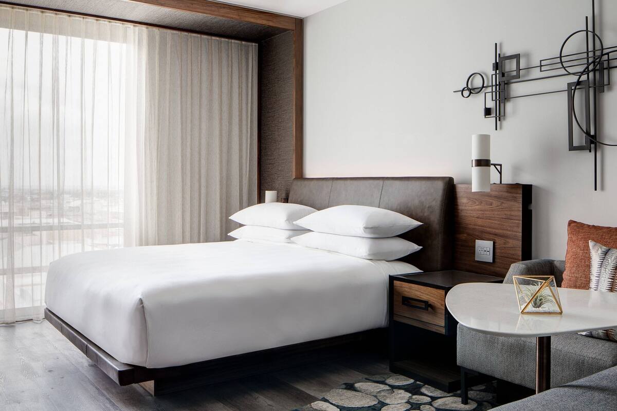 A modern hotel room with a large bed, a bedside table, a small round table, and a wall-mounted light fixture.