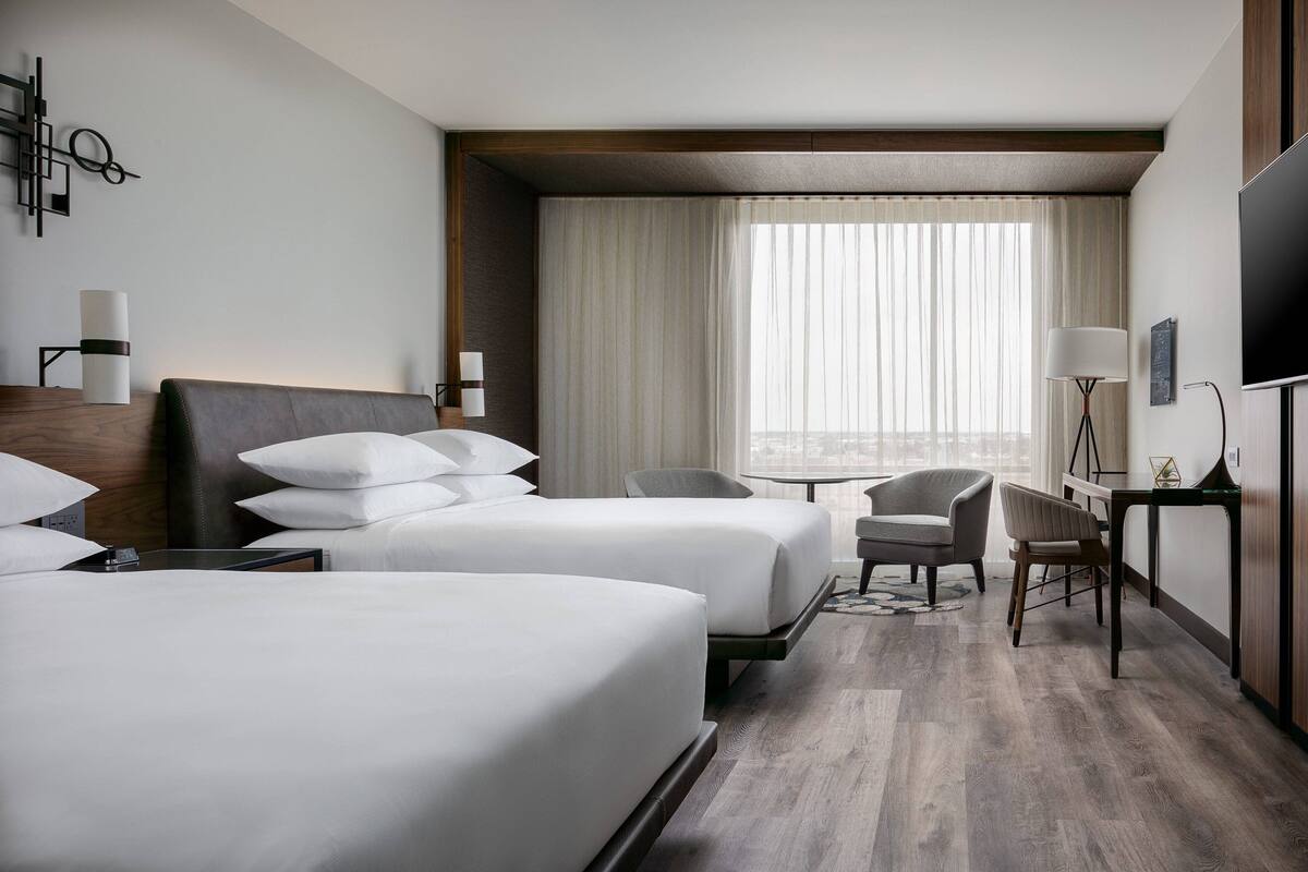 A hotel room with two beds, a desk, a chair, a TV, and a large window with curtains.