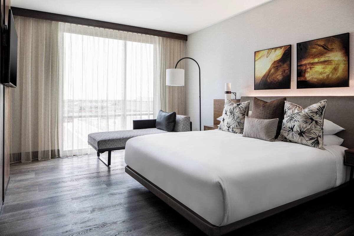 A modern hotel room with a large bed, a bench, a bedside table with a lamp, and artwork on the wall.