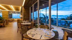 A modern dining area with round tables, wicker chairs, and large windows offering a view of the ocean.