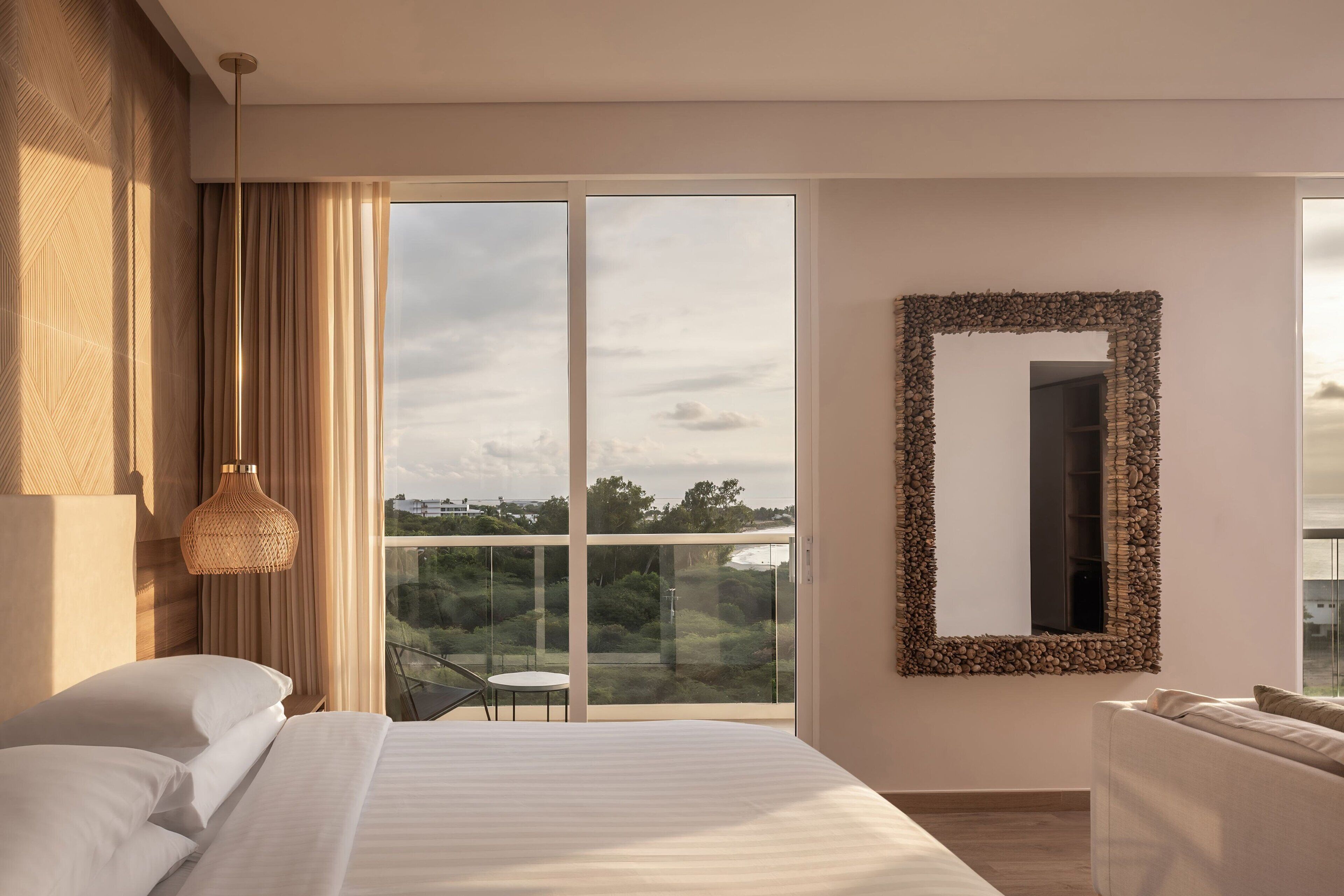 A modern bedroom with a large bed, a balcony with a glass door, a woven light fixture, and a unique stone-framed mirror.