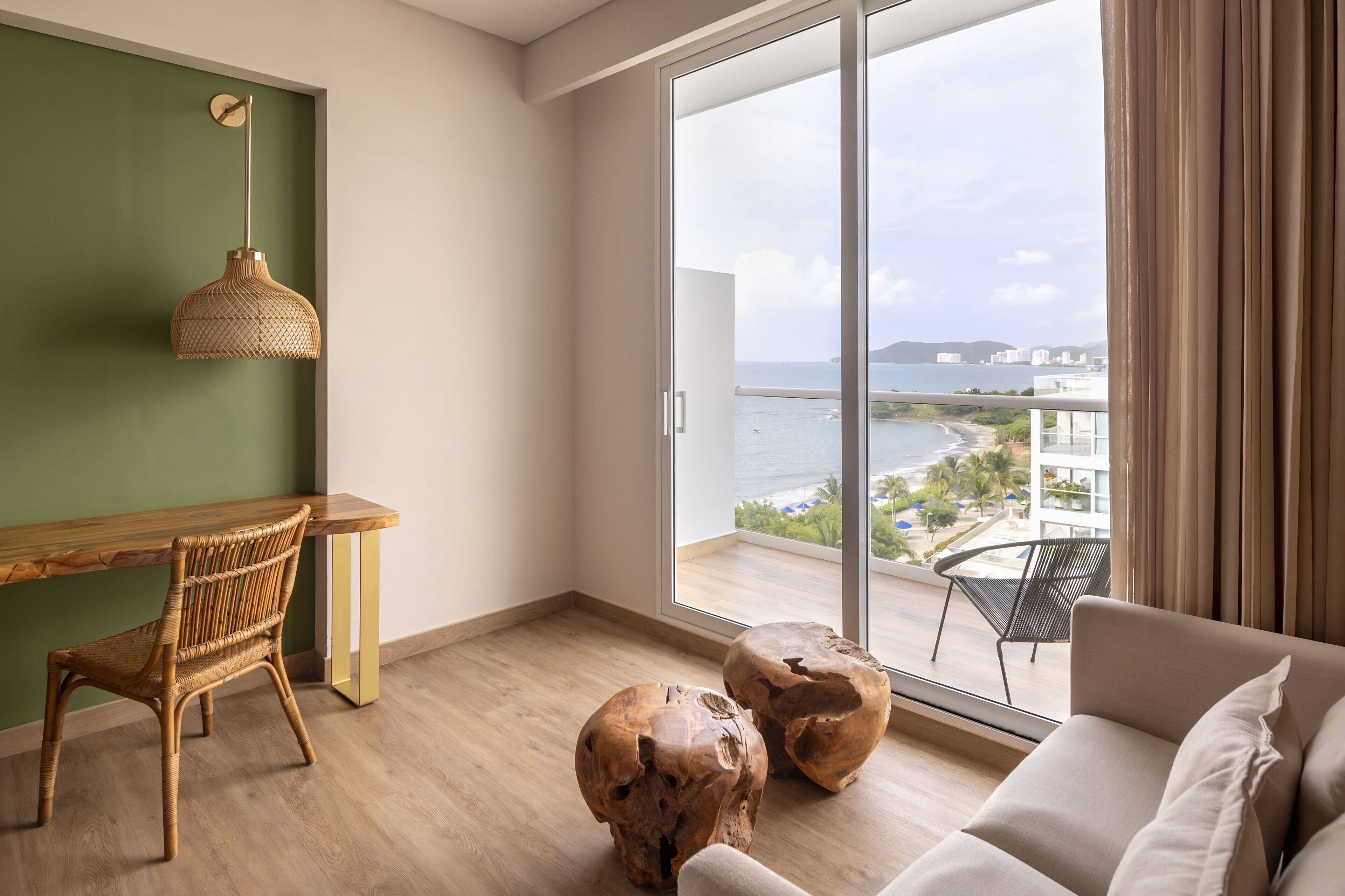 A modern room with a wooden desk, a wicker chair, a sofa, and a large sliding glass door leading to a balcony with a view of the sea and buildings.