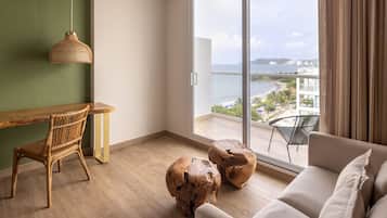 A modern room with a wooden desk, a wicker chair, a sofa, and a large sliding glass door leading to a balcony with a view of the sea and buildings.