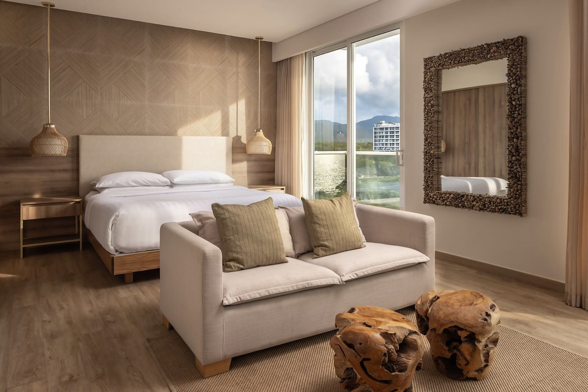 Een moderne hotelkamer met een groot bed, een bank, een houten salontafel en een groot raam met uitzicht over de stad.