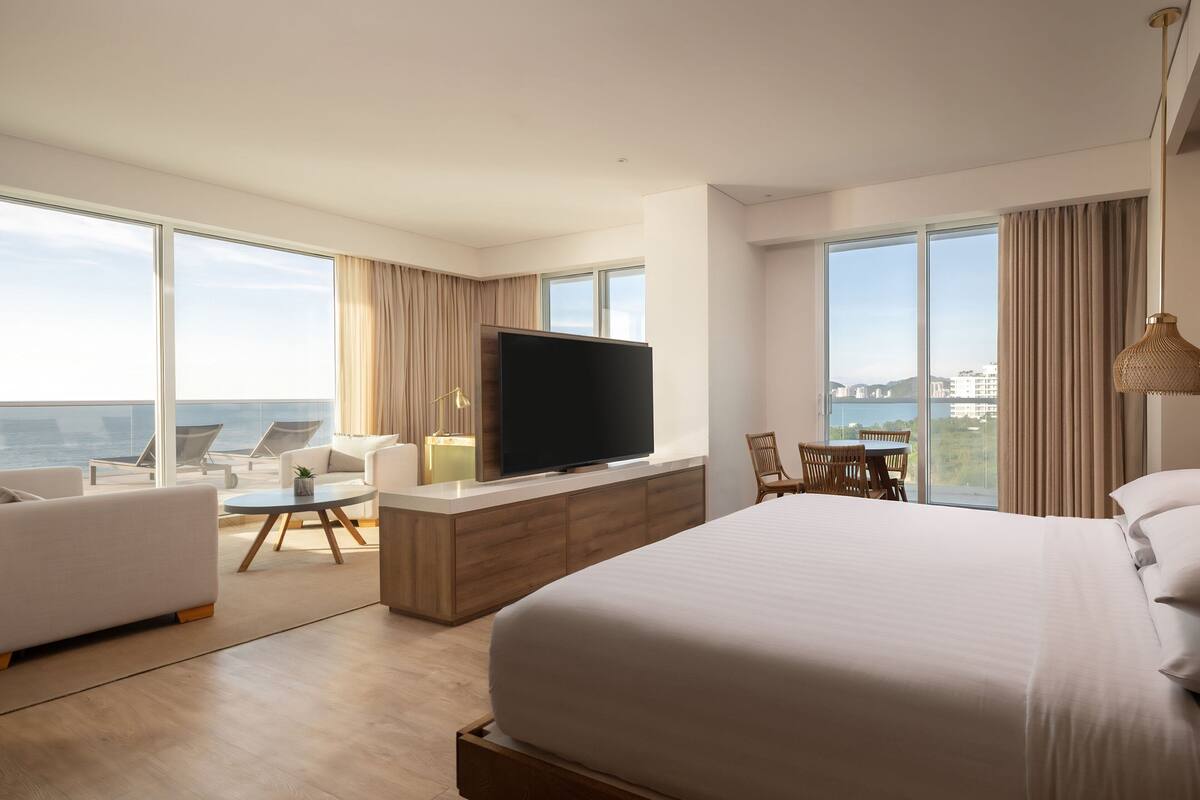 Een moderne hotelkamer met een groot bed, een flatscreen televisie, een zithoek met een tafel, en grote ramen met uitzicht op de stad.