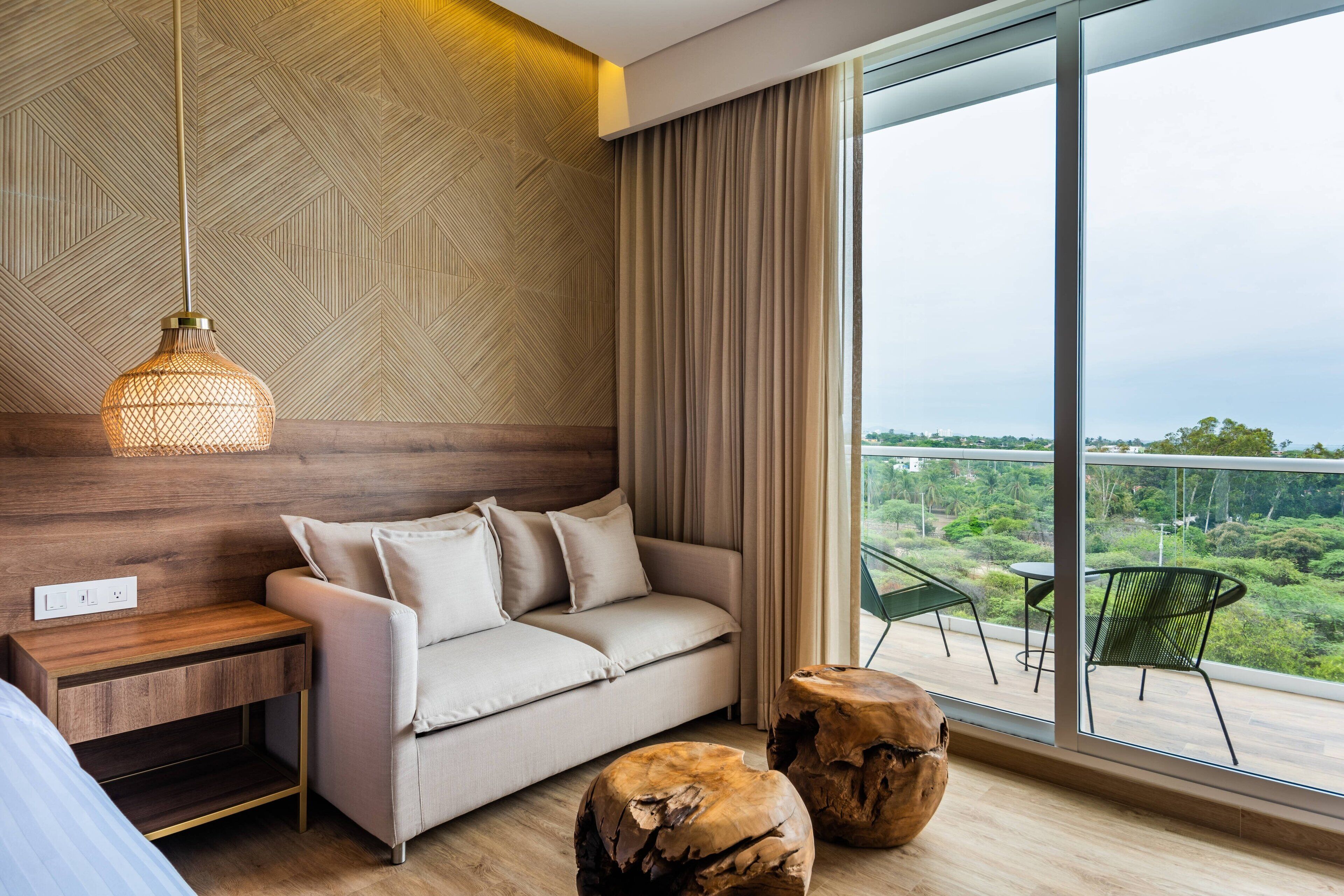A modern room with a sofa, a wooden side table, a hanging light fixture, and a view of greenery through large windows.