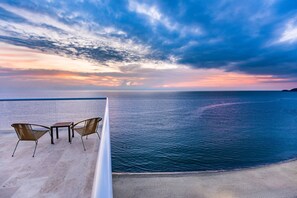 A balcony with two chairs and a table overlooking a calm sea at sunset.